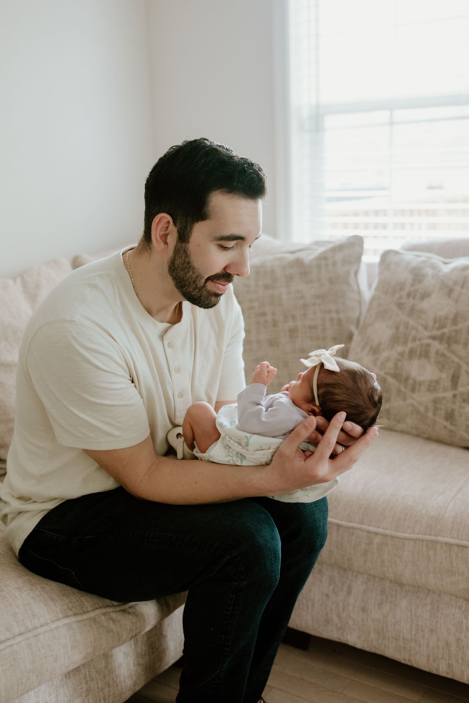 A father tenderly holds his newborn baby girl with a bow headband while sitting on a beige sofa.