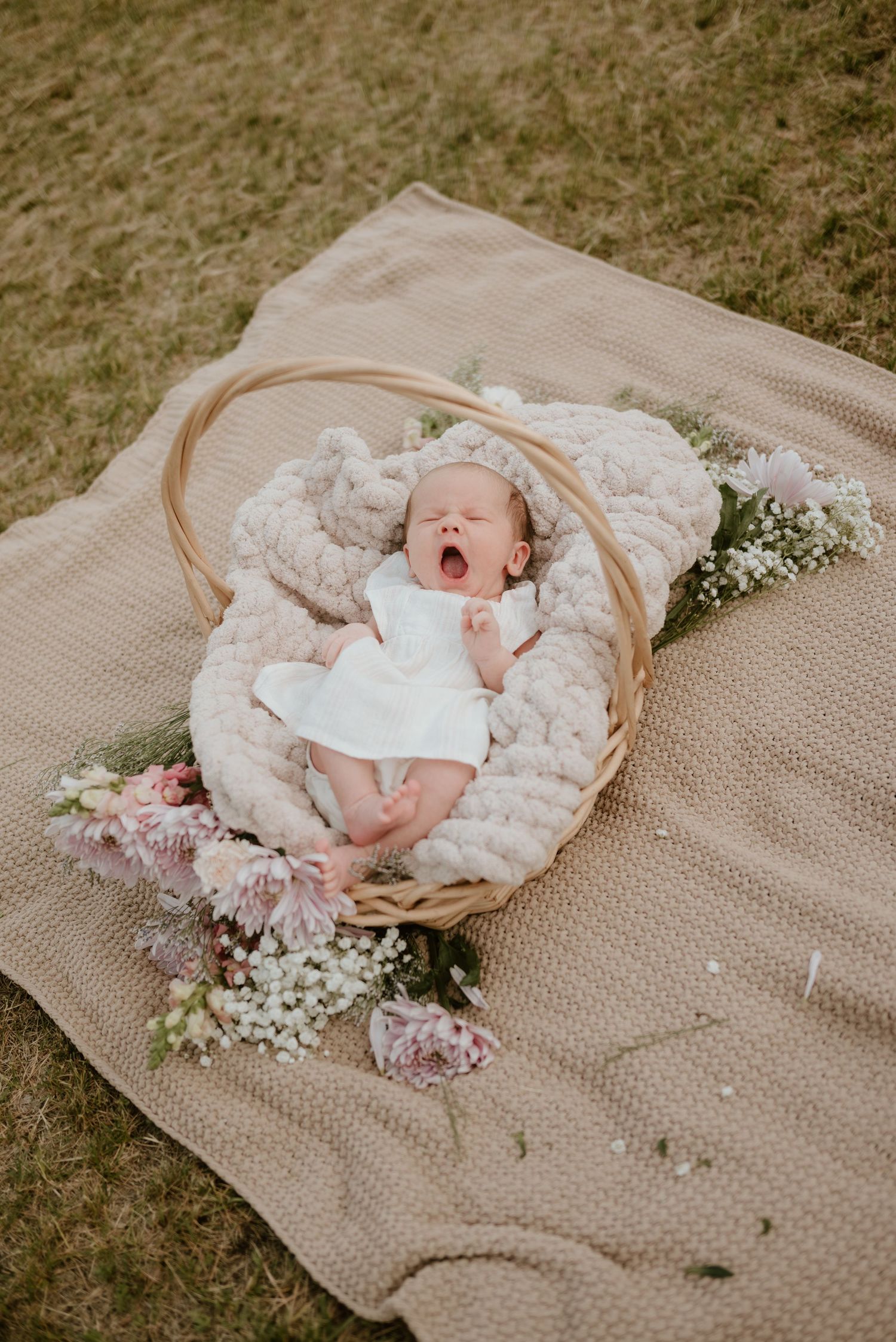 Newborn baby yawning in a wicker basket with soft blanket and pink flowers on a beige blanket outdoors.
