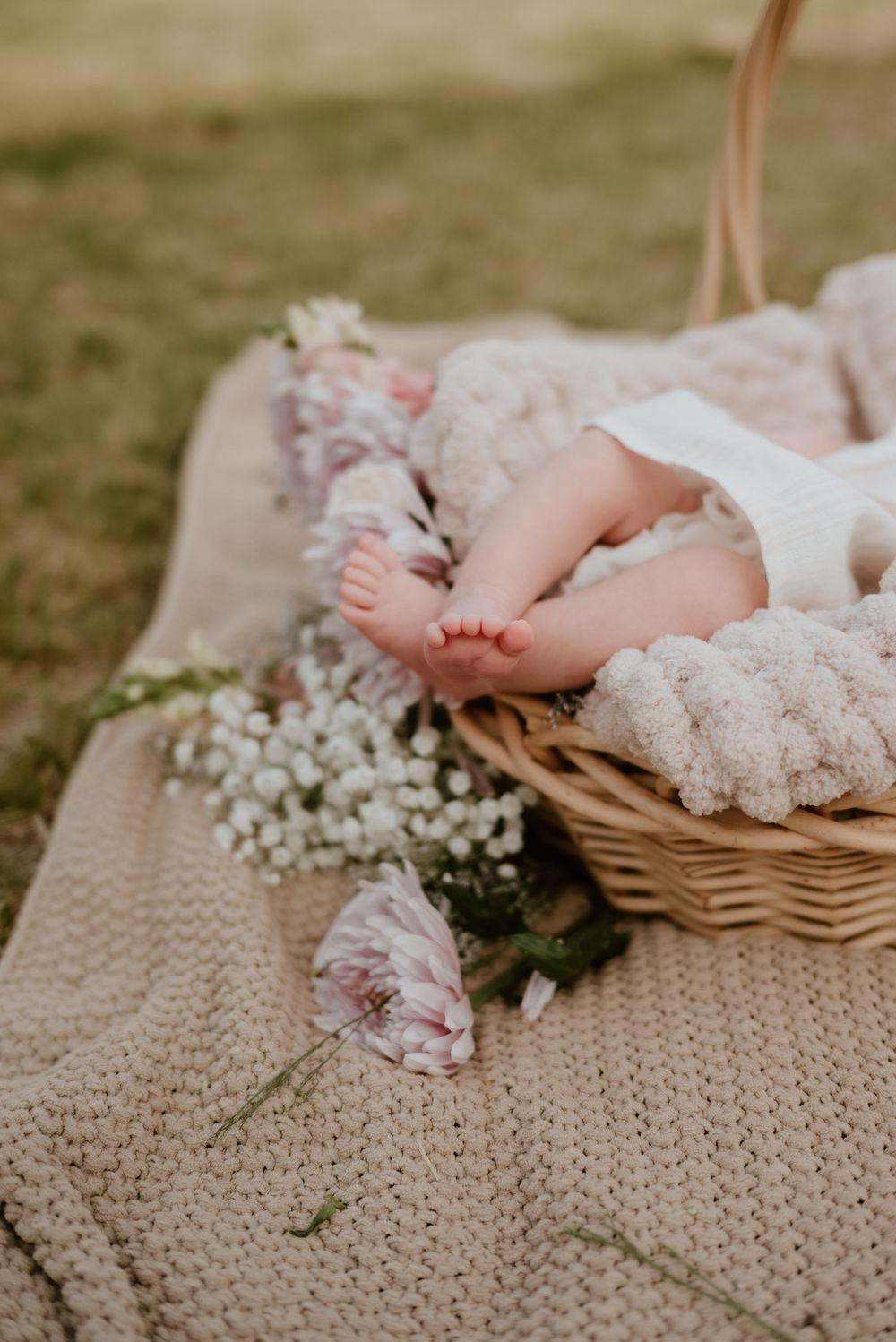 Tiny baby feet peeking out of a wicker basket on a knit blanket surrounded by delicate white and pink flowers outdoors.