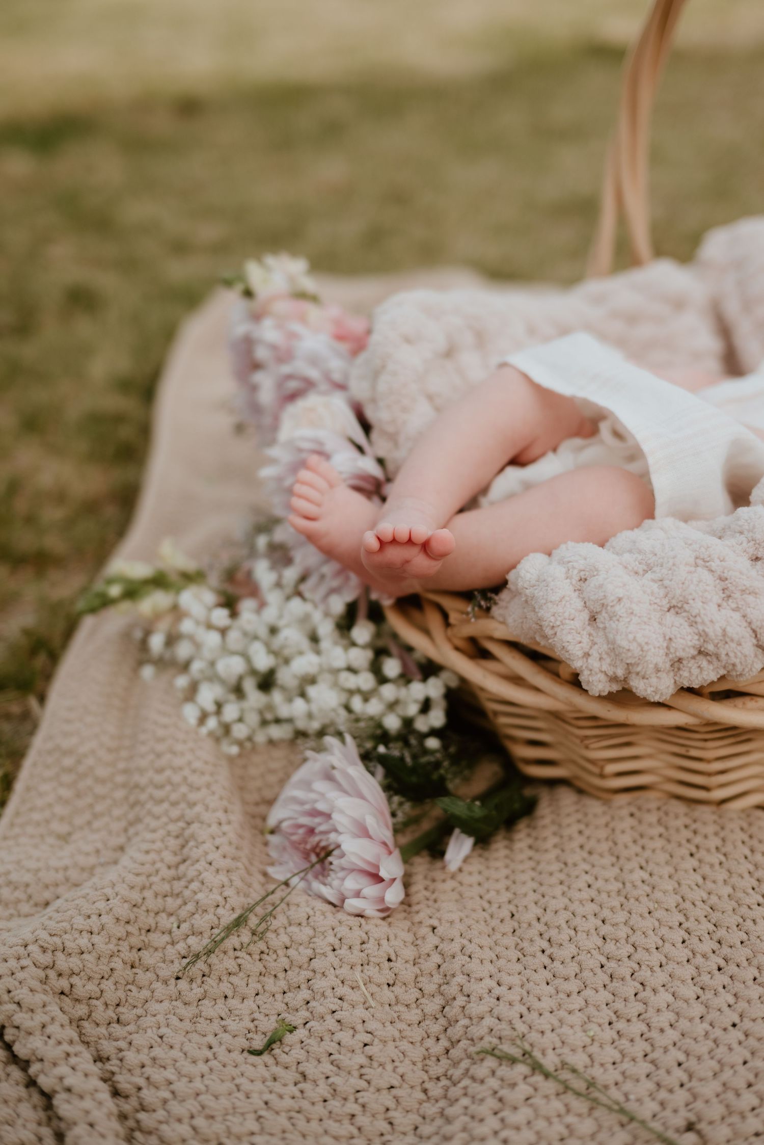 Tiny baby feet peeking out of a wicker basket on a knit blanket surrounded by delicate white and pink flowers outdoors.