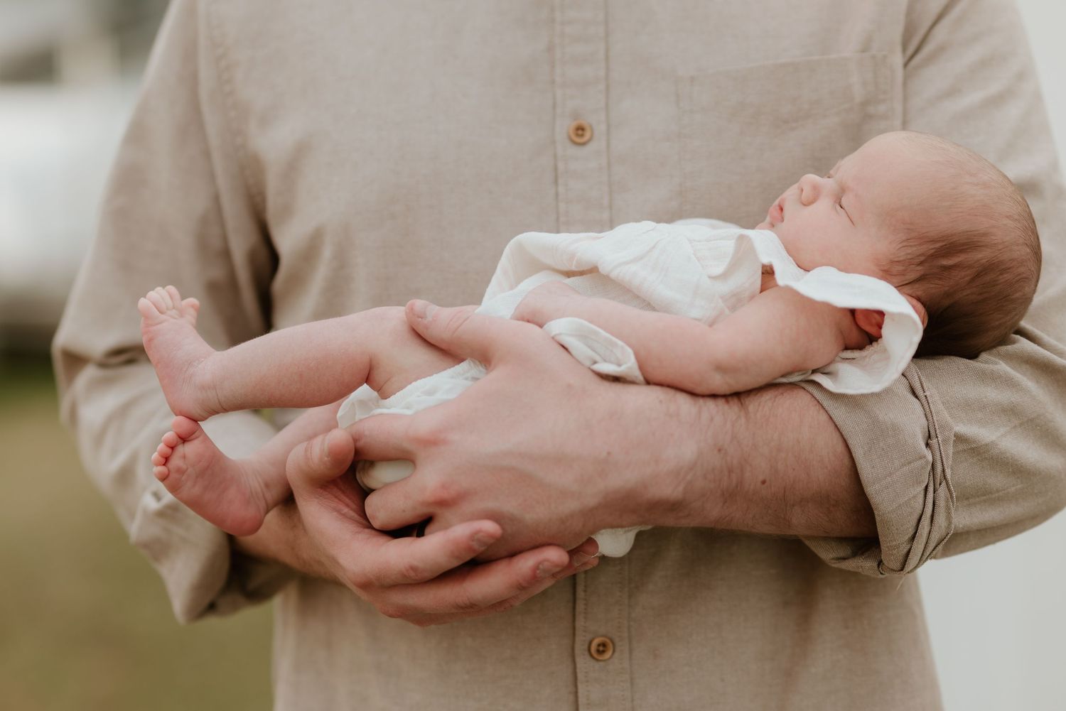 A father cradles a sleeping newborn baby in his hands, wearing a beige linen shirt outdoors.