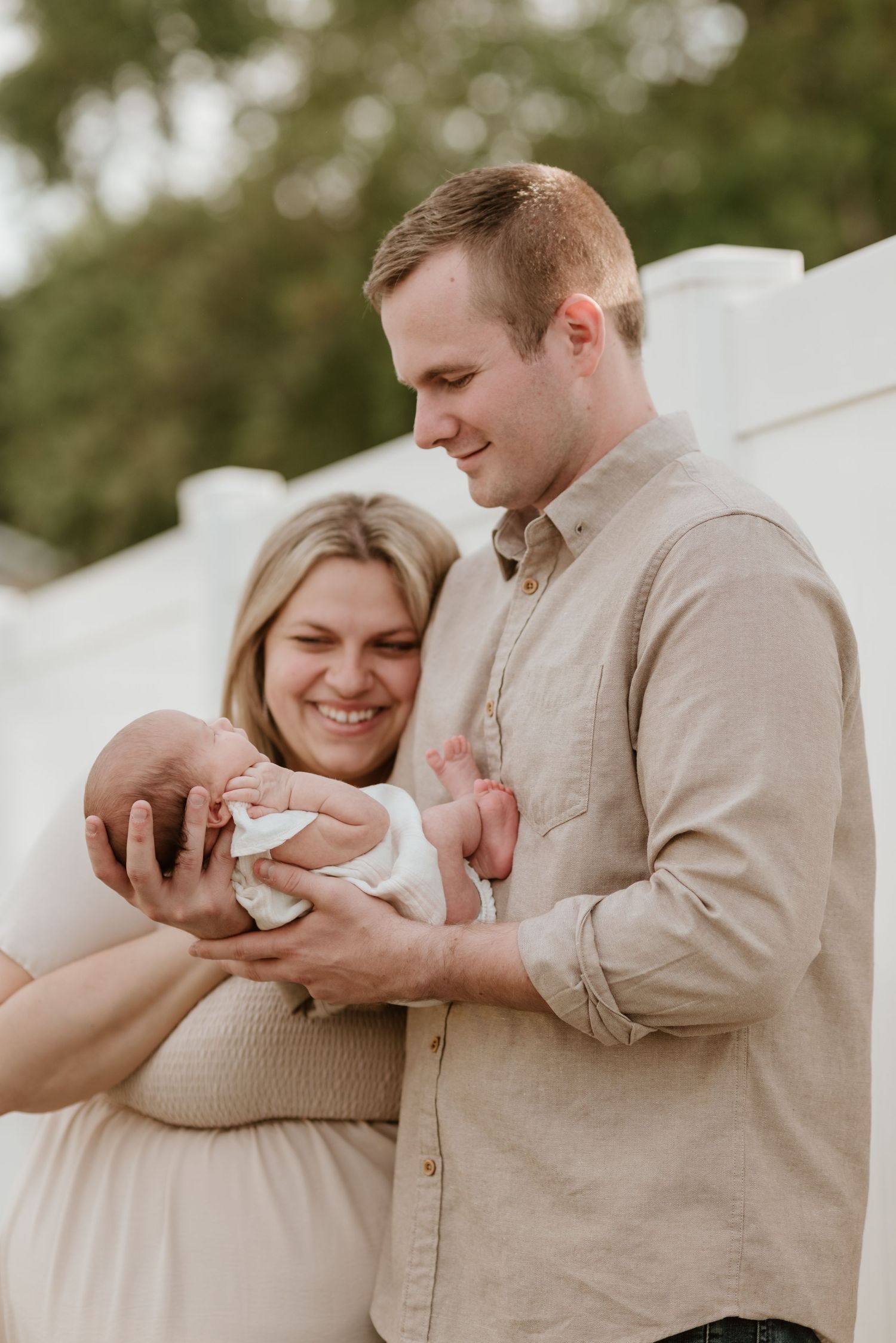 Smiling couple admiring their newborn baby outdoors, dressed in neutral tones against a white backdrop.