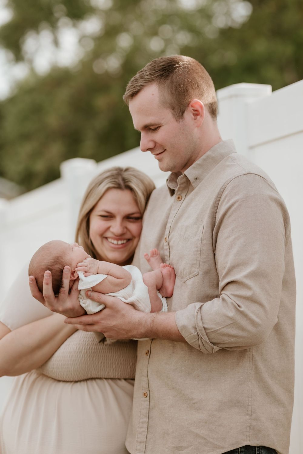 Smiling couple admiring their newborn baby outdoors, dressed in neutral tones against a white backdrop.