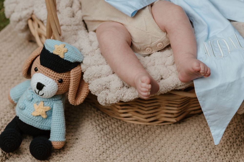 Newborn baby feet in a wicker basket beside a crocheted police bear toy on a beige knit blanket.