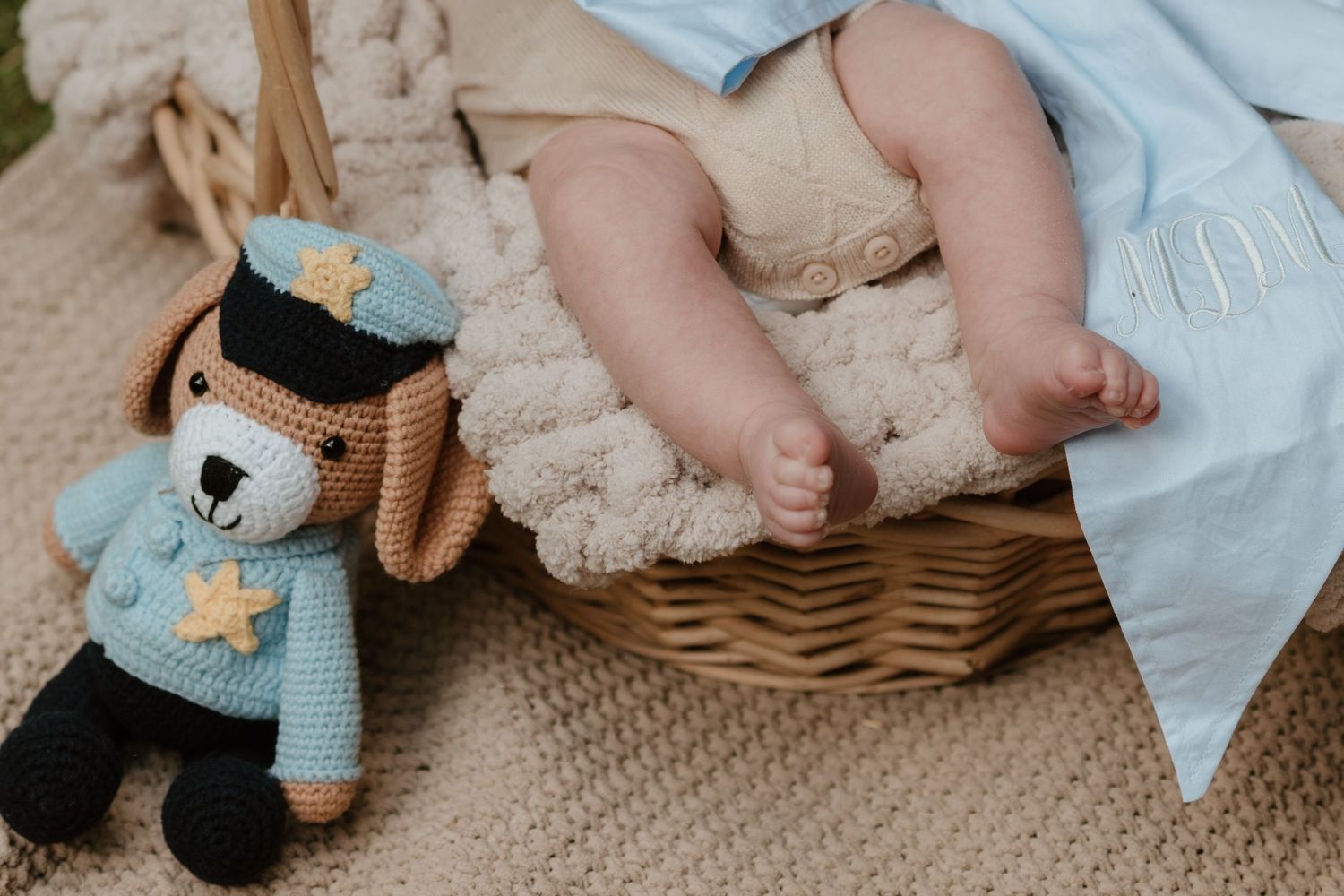 Newborn baby feet in a wicker basket beside a crocheted police bear toy on a beige knit blanket.