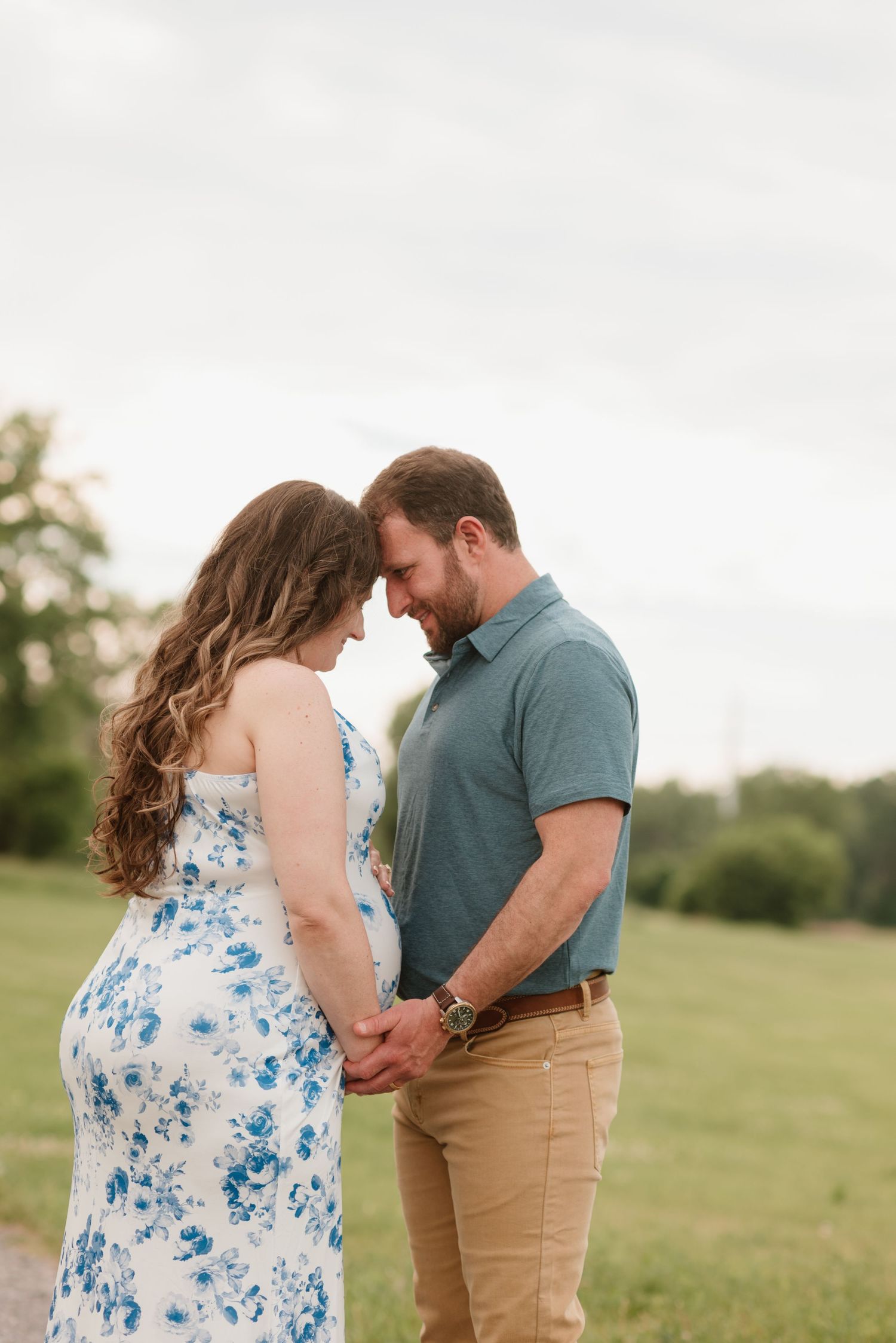 Pregnant couple holding hands and touching foreheads in a green field, woman in floral dress, man in teal polo.