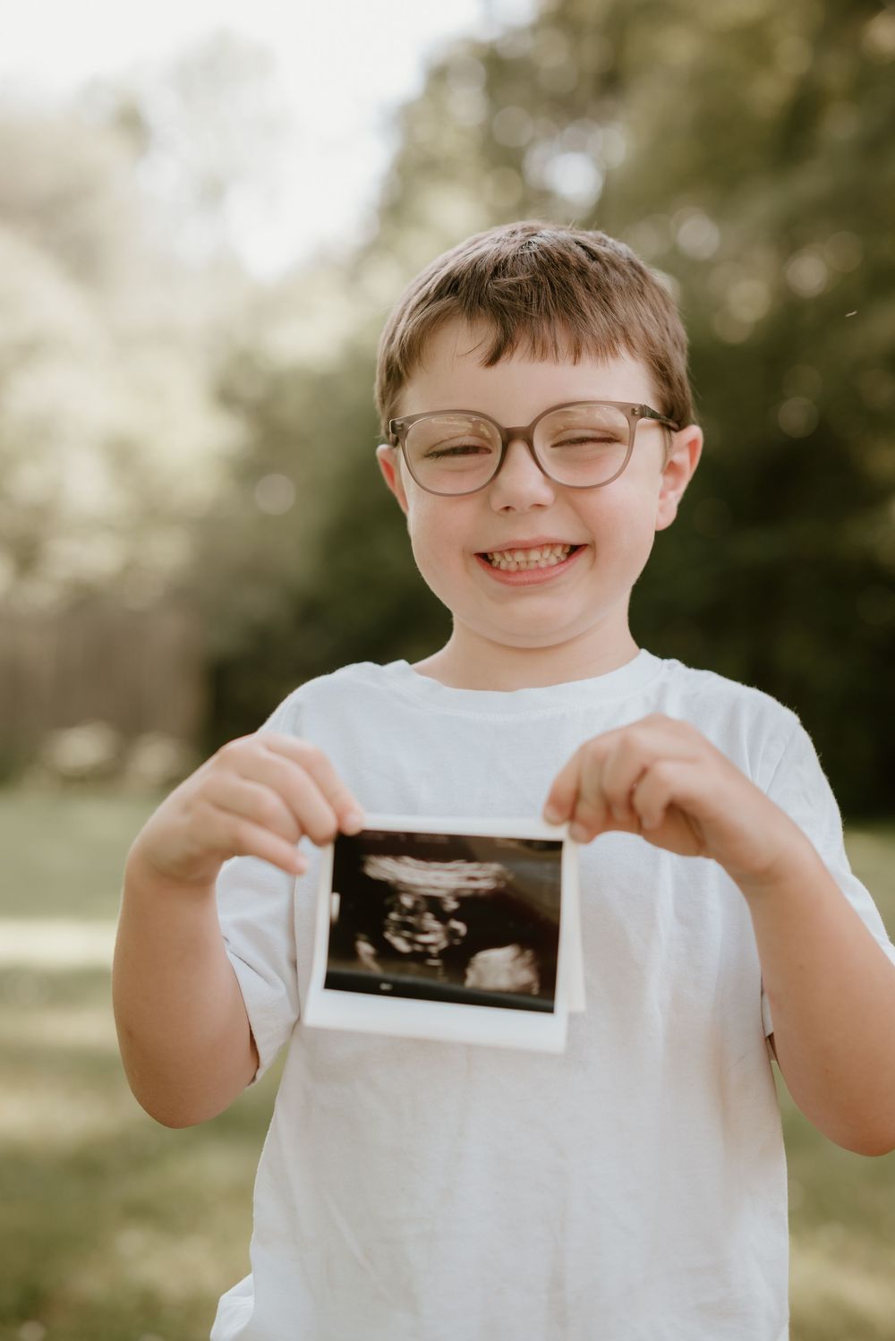 Smiling boy with glasses proudly holding ultrasound photo outdoors.