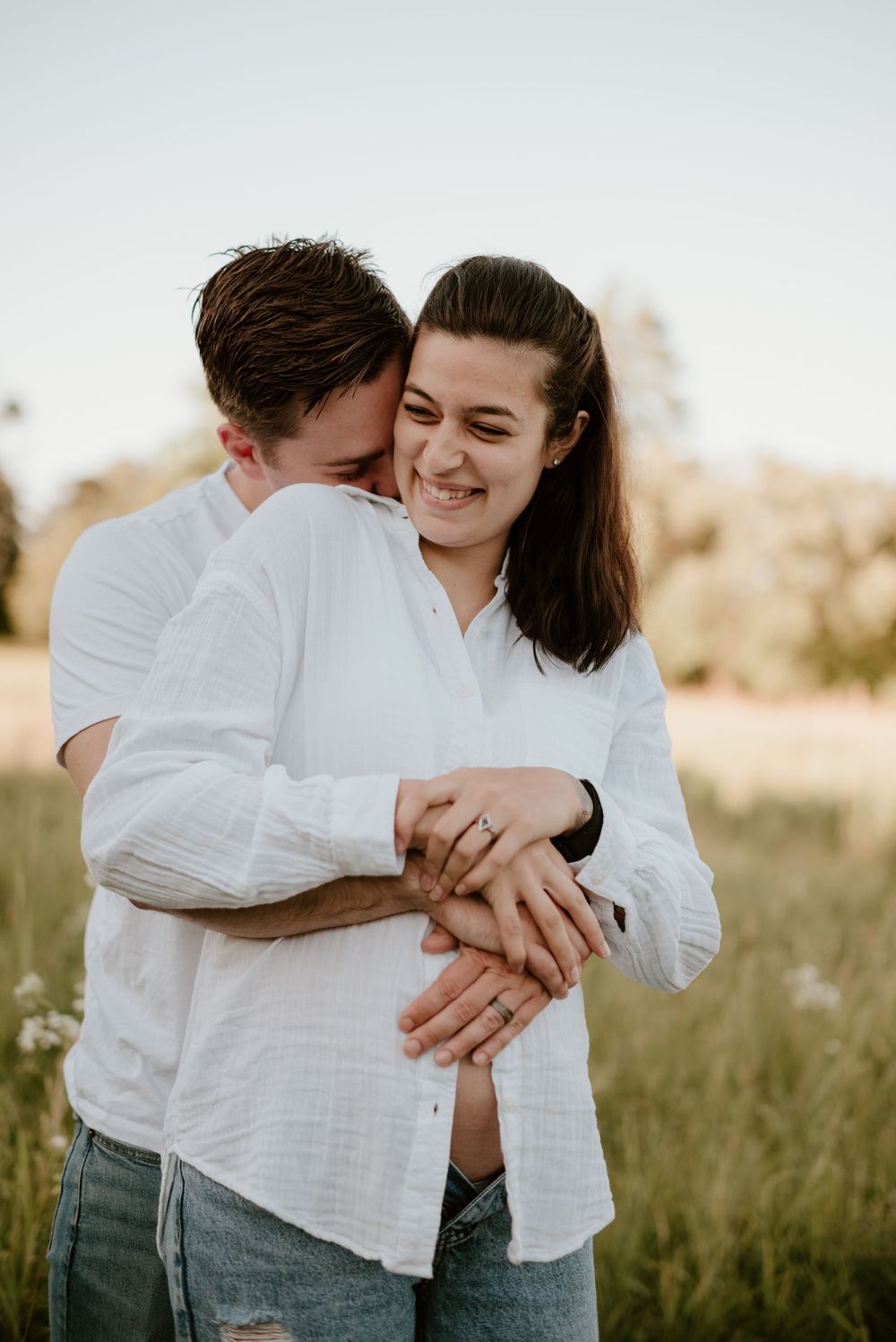 Happy couple embracing in a sunny field, woman smiling as man hugs her from behind showing engagement ring.