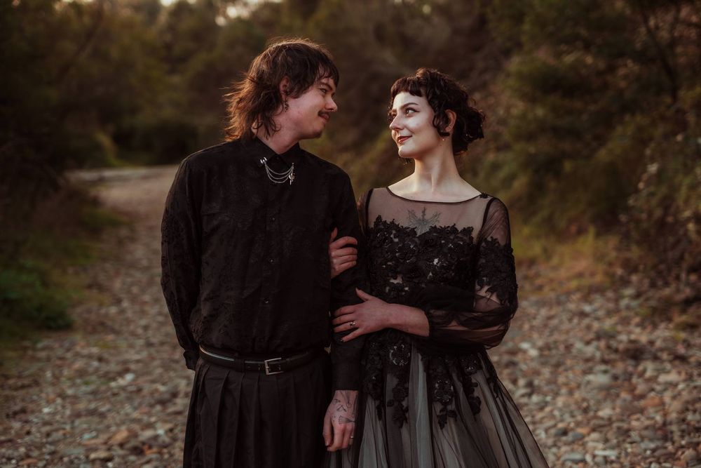 Couple in matching black gothic wedding attire walking together on a scenic outdoor path at sunset.