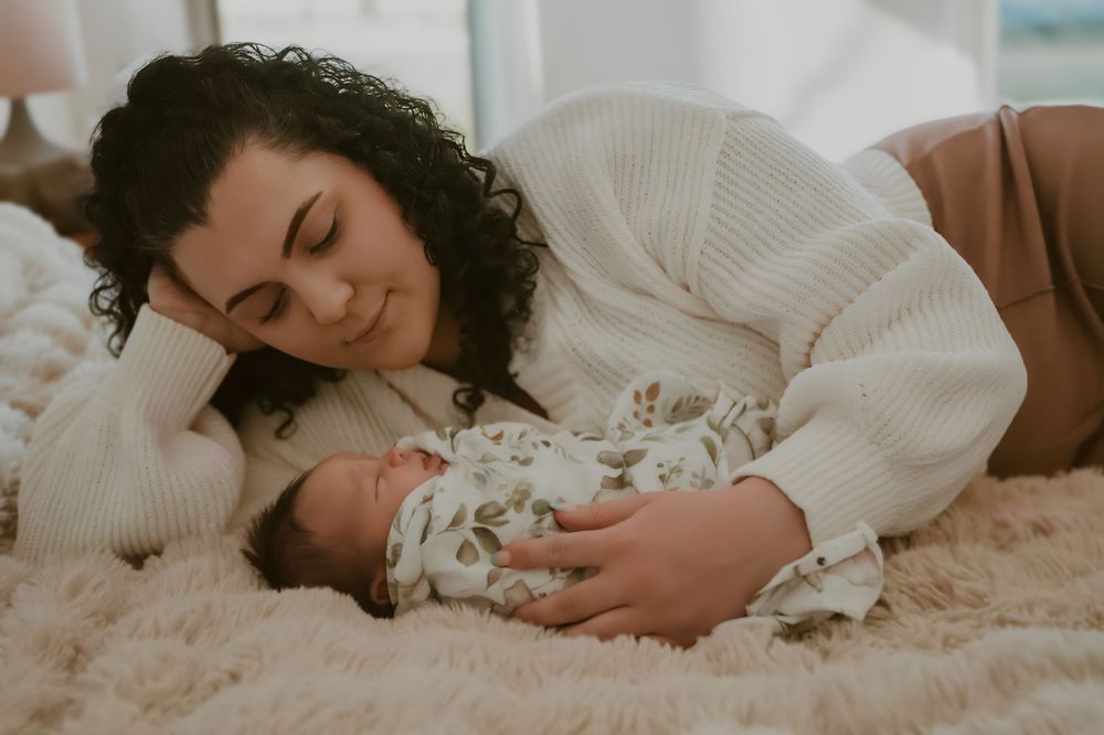 Mother lying on a fluffy rug smiling lovingly at her newborn baby in a floral wrap.