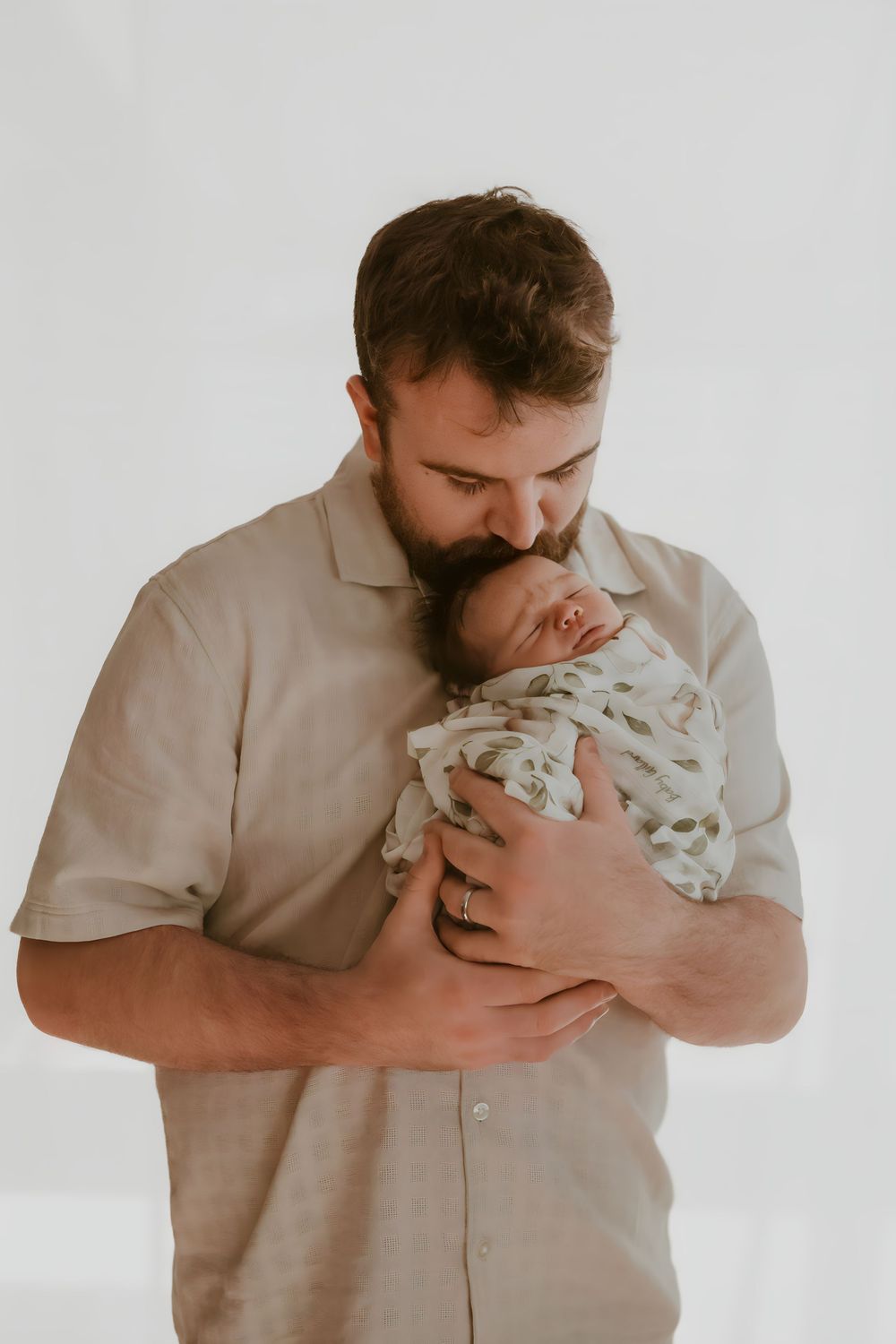 Bearded father gently kisses his swaddled newborn baby against a white background.