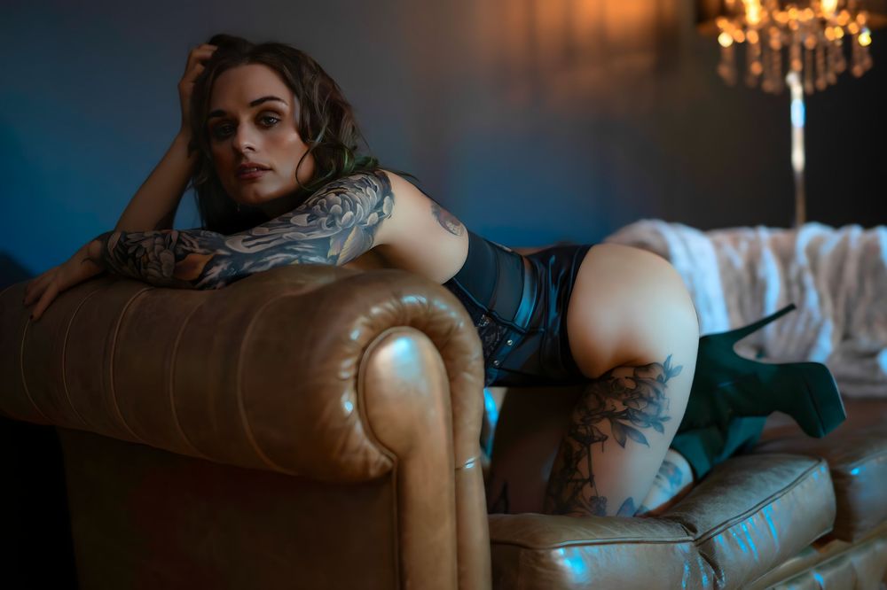 Tattooed woman in lingerie posing on a leather sofa in moody blue lighting.
