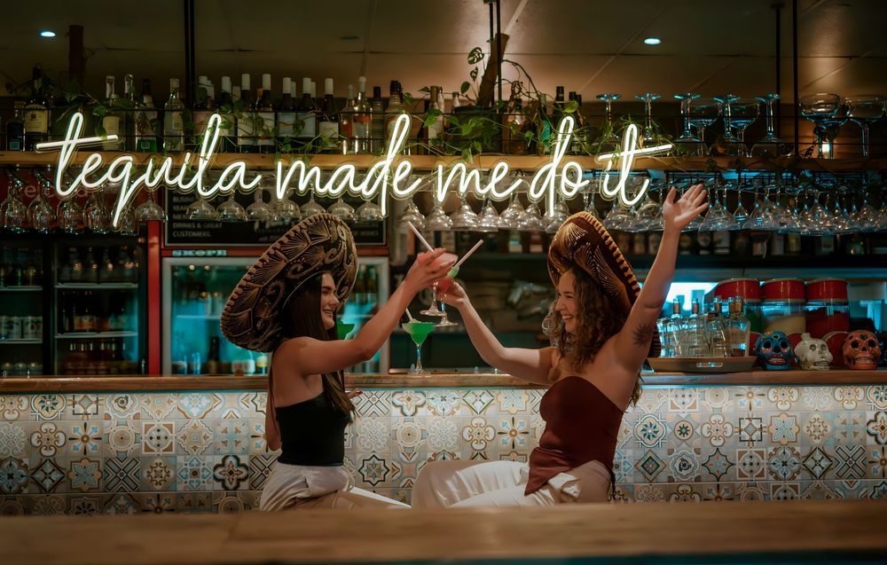 Two women in sombreros toast drinks at a bar under a tequila made me do it neon sign.