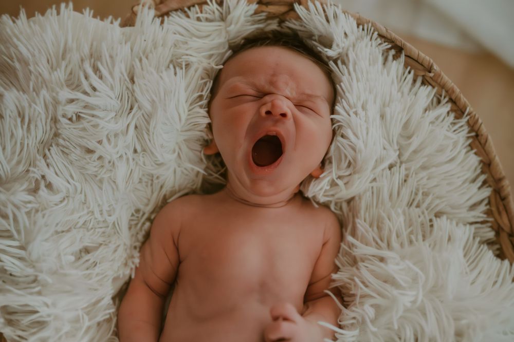 Newborn baby yawning while lying on a fluffy white blanket in a round basket.