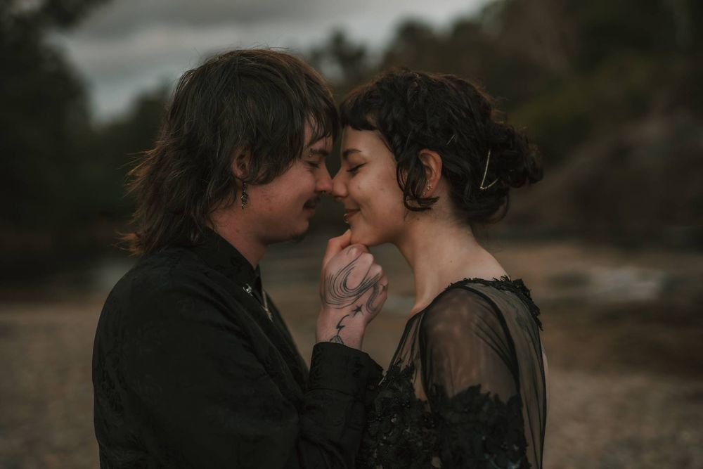 Romantic couple in dark attire sharing an intimate moment outdoors, touching foreheads and holding hands.
