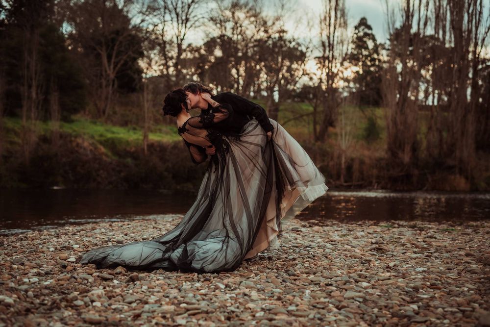 Romantic dip pose by a river, groom in black holding bride in elegant dark wedding dress on pebble shore.