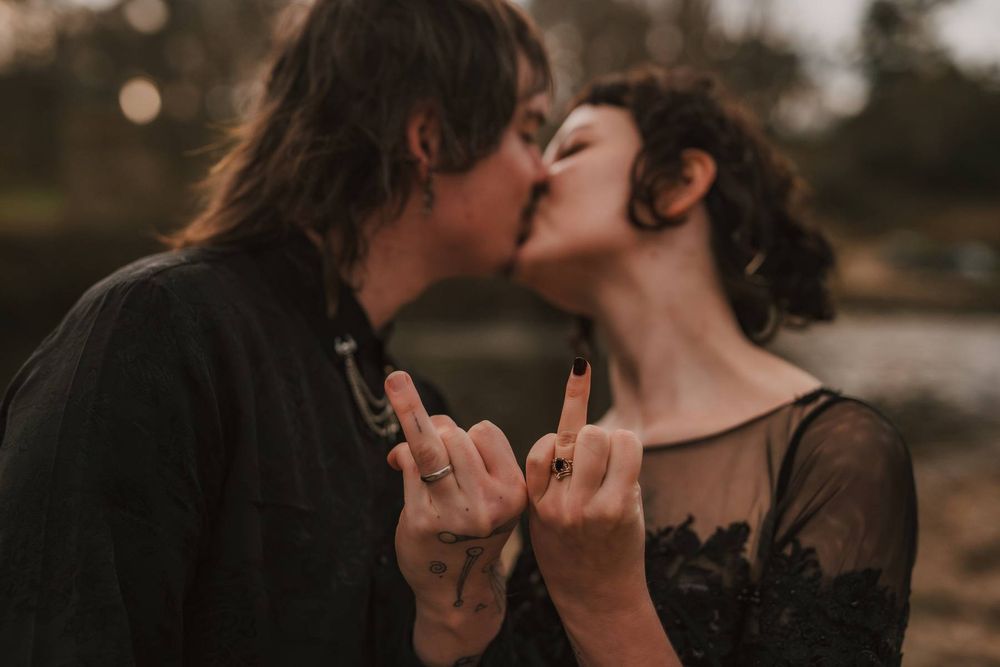 Edgy couple kissing while showing rock on hand gestures wearing black lace and linen wedding outfits.