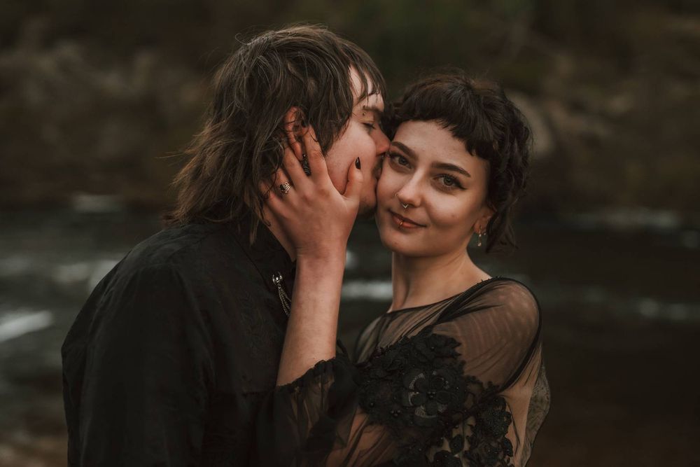 A couple in dark romantic attire shares an intimate moment near a misty river, the woman smiling softly.