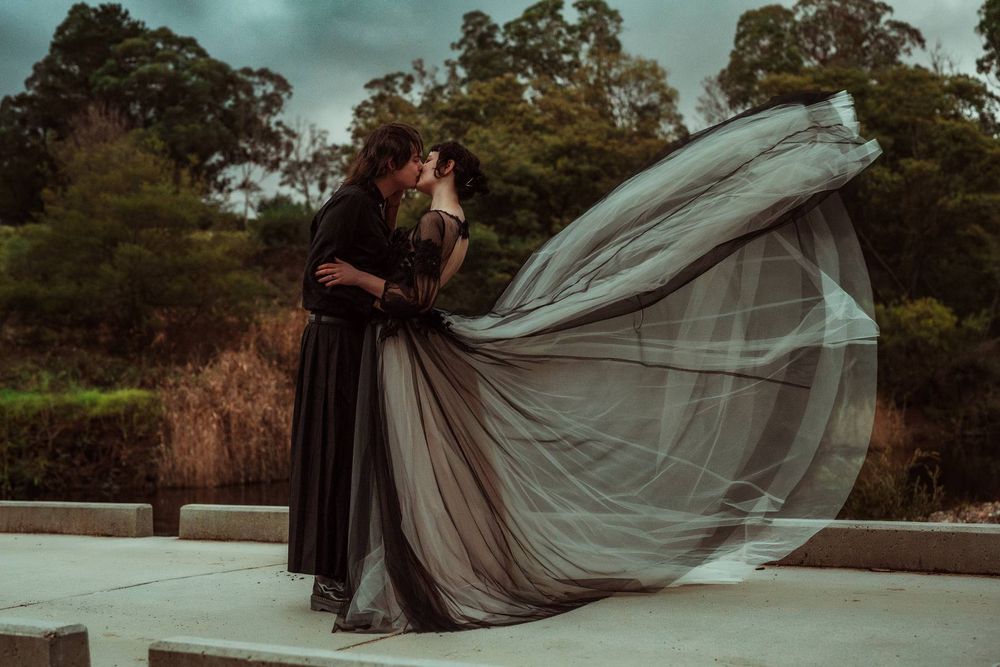 Couple kissing in dramatic dark wedding attire with flowing grey tulle gown billowing in wind outdoors.