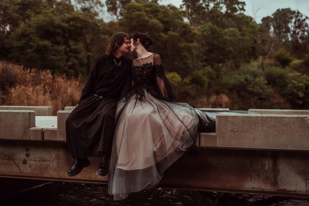 A couple in gothic wedding attire share a romantic moment sitting on a concrete bridge surrounded by lush greenery.