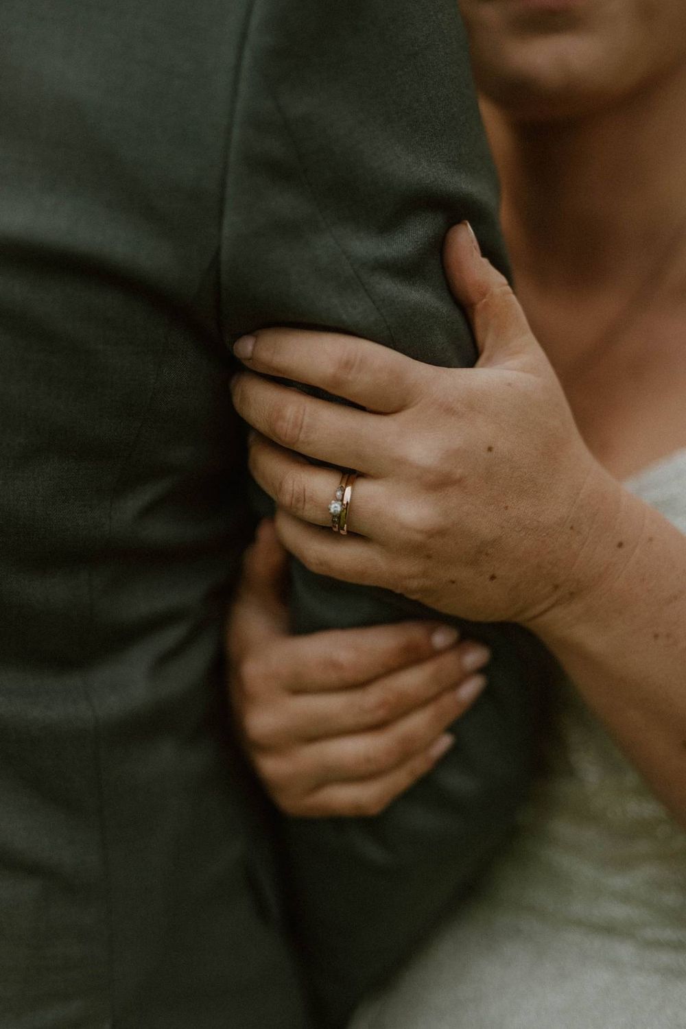 Bride with wedding ring gently holds grooms gray suit jacket in close-up detail shot.