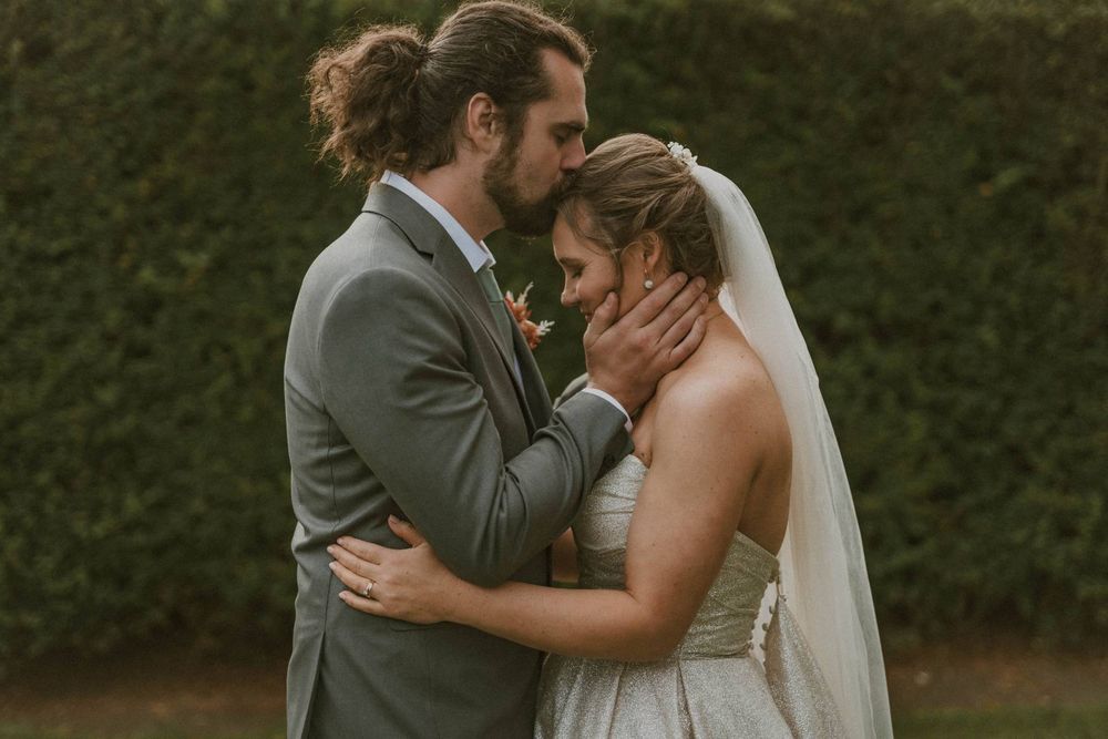 Groom gently kisses bride on forehead while embracing her outdoors at garden wedding.