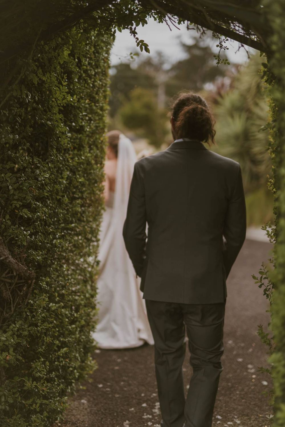 Groom walks toward bride in white gown through lush green garden archway at outdoor wedding.