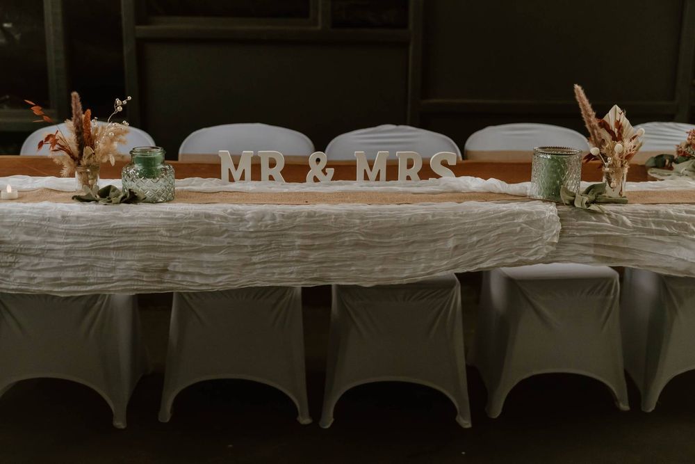Wedding reception head table decorated with MR & MRS sign, white linens, and dried floral arrangements.