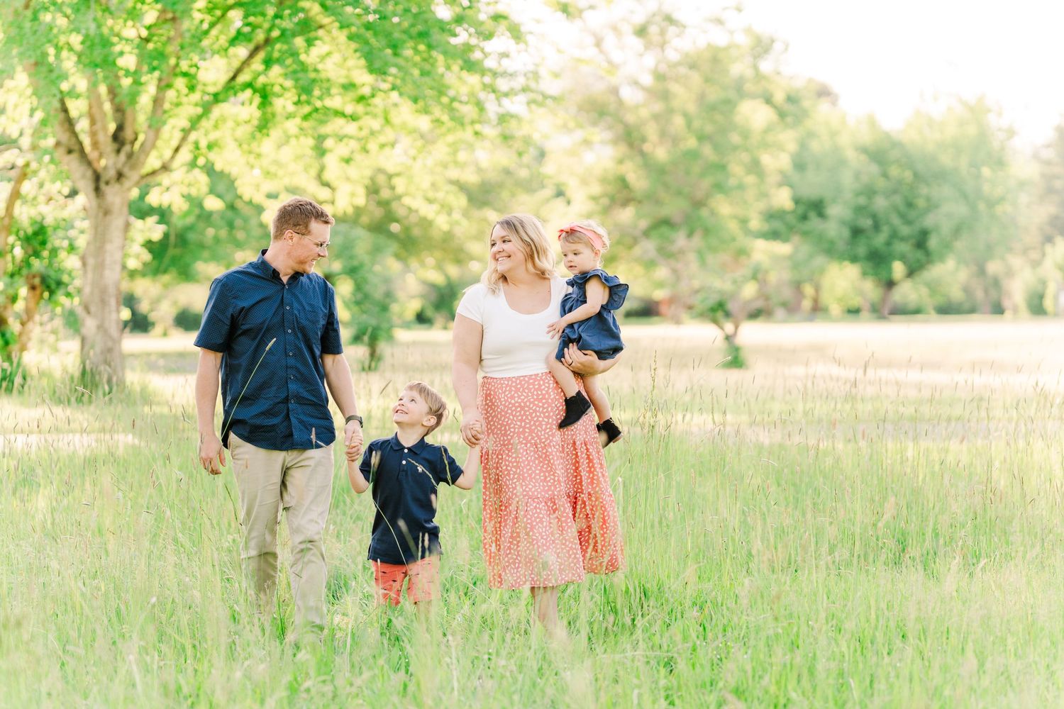 Young family of four walking together through a lush green meadow on a sunny day.