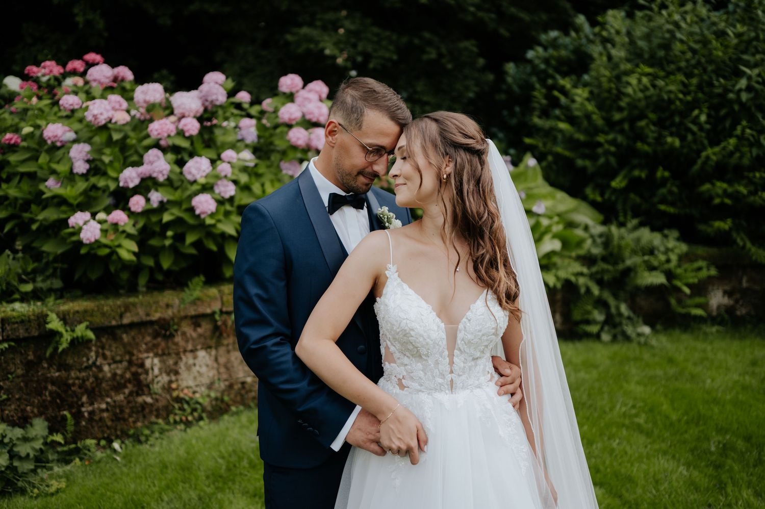 Romantisches Hochzeitspaar umarmt sich in einem üppigen Garten mit blühenden rosa Rosen im Hintergrund.