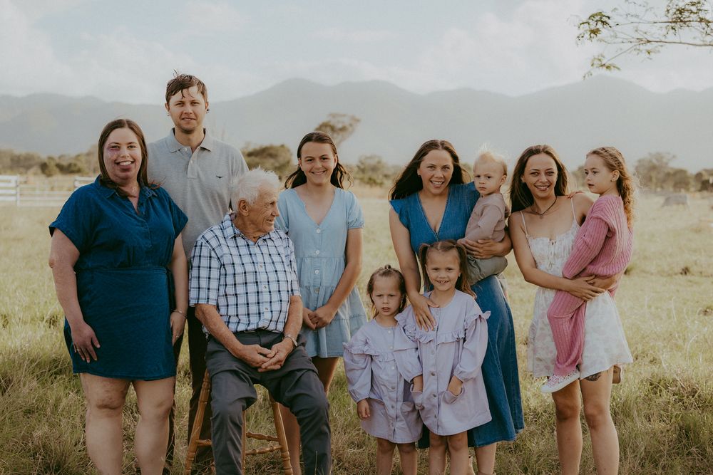 Multigenerational family group portrait with grandfather seated in sunny rural field.