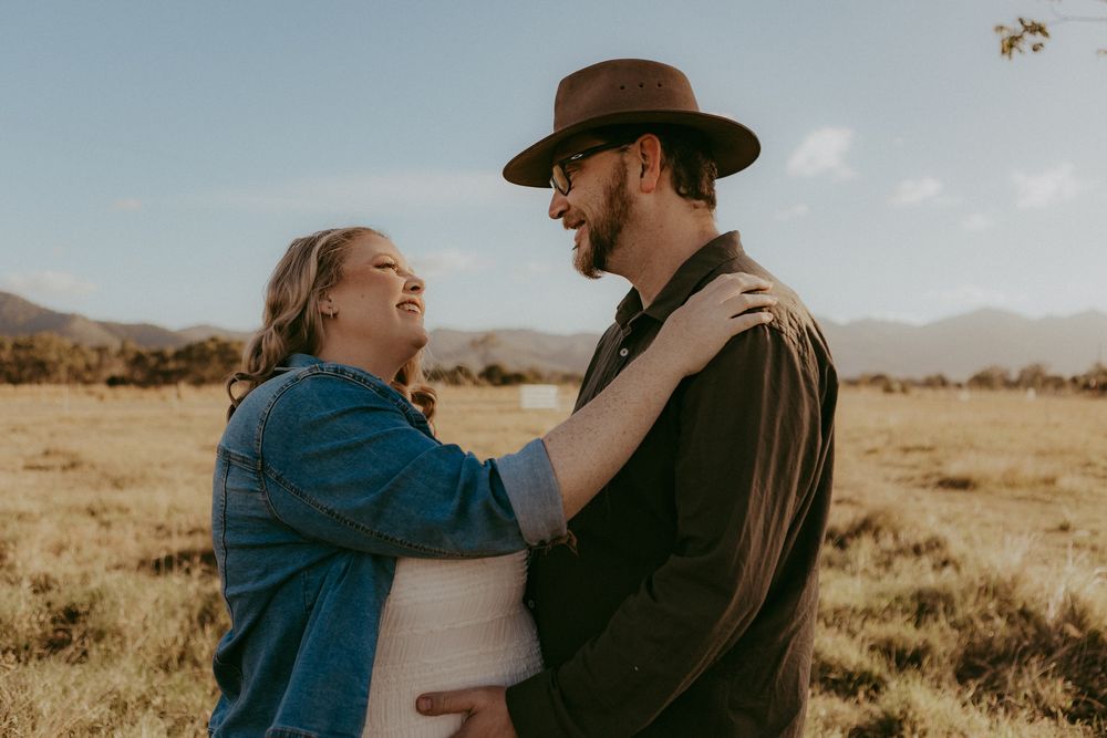 Smiling pregnant couple facing each other in a bright open field with mountains in the background.