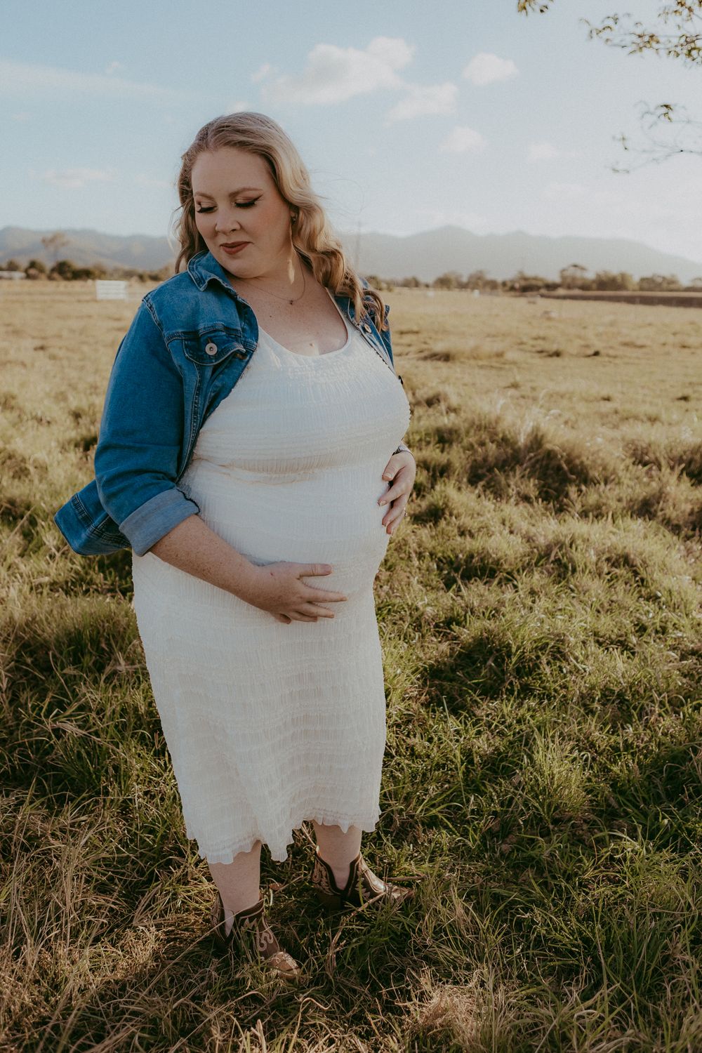 Pregnant woman in white dress and denim jacket cradling baby bump in golden hour field.