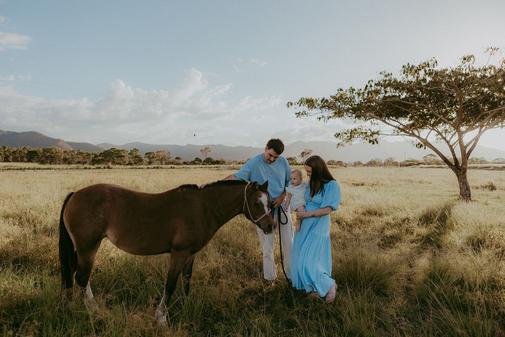 Young family in blue outfits interacting with brown horse in scenic rural field.