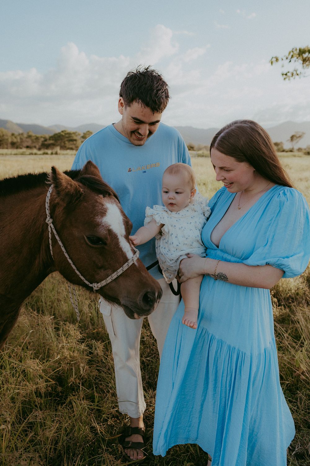Young family with baby meets a brown horse in a sunny field with mountain backdrop.