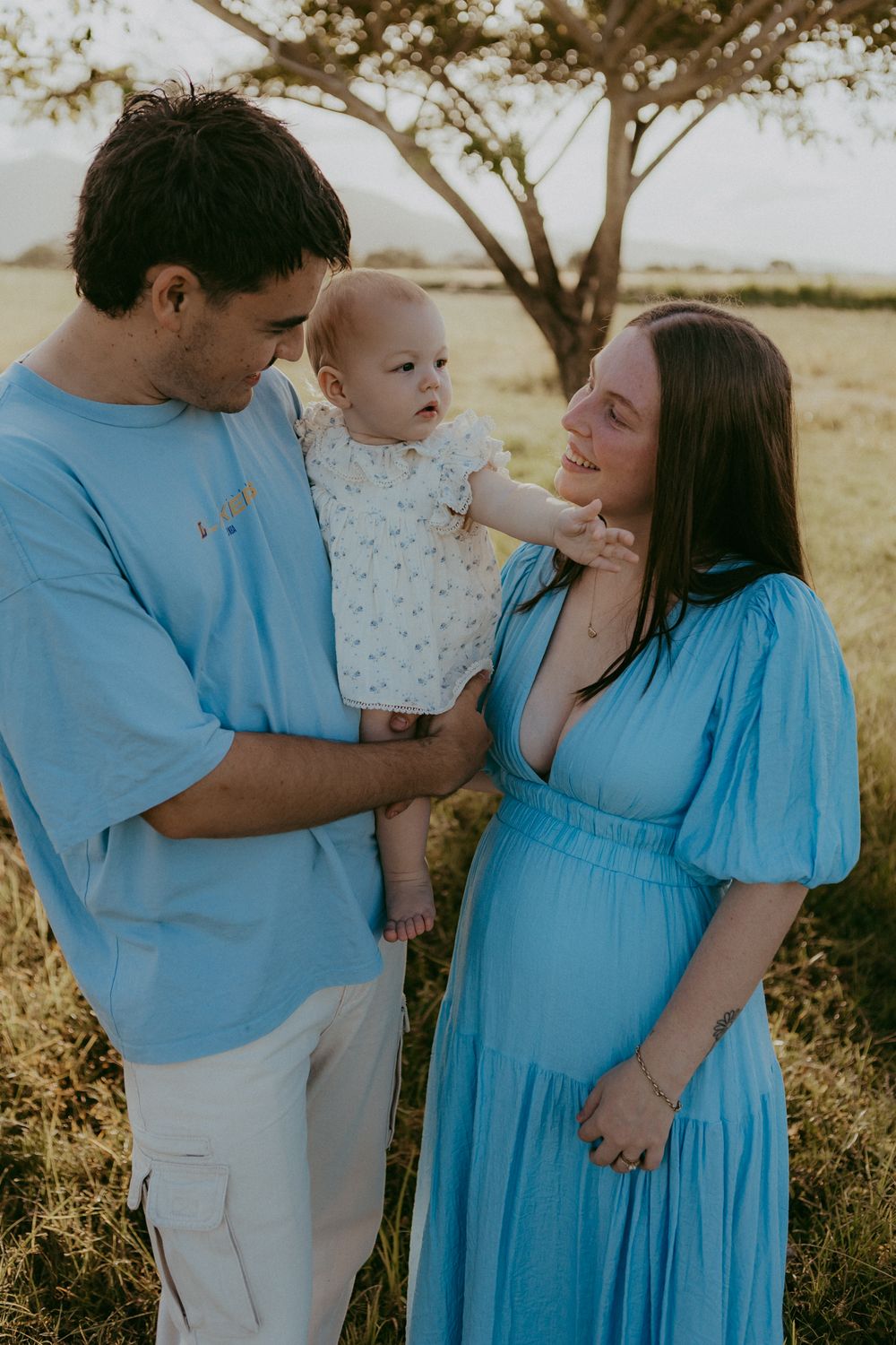 Young couple in blue outfits holding infant baby together under tree in golden field.