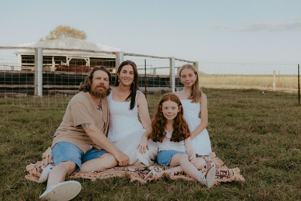 Family of four sitting on blanket in farm field with cattle pen in background.