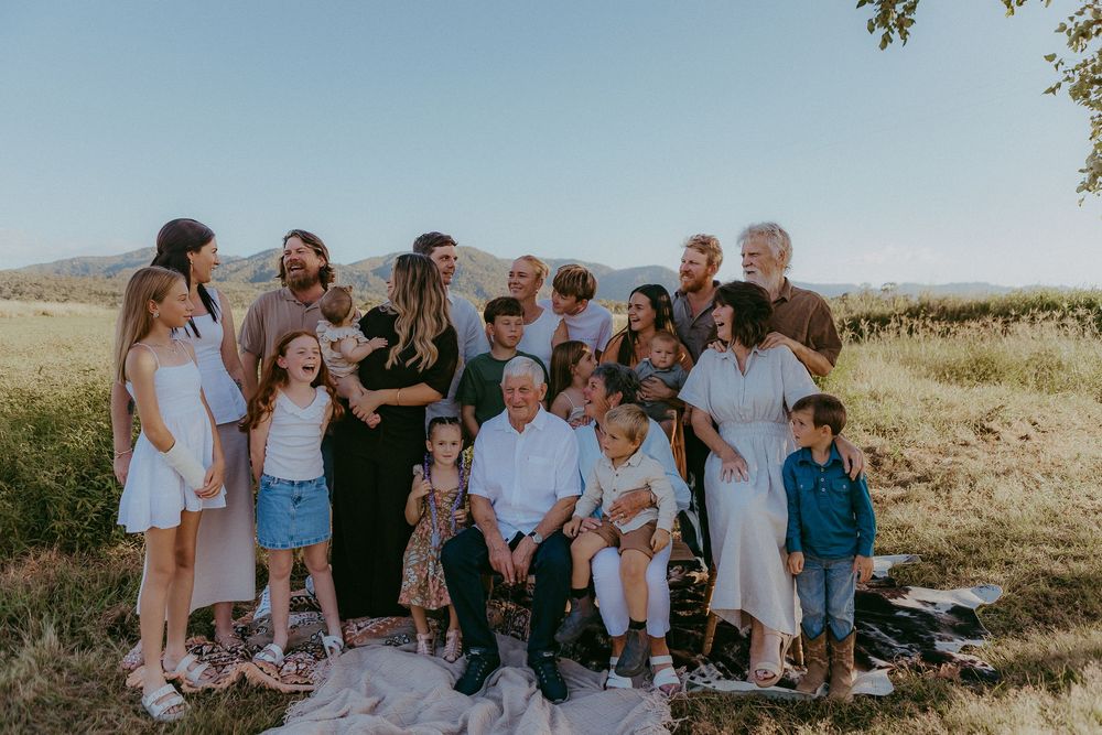 Large extended family gathering outdoors in a sunny field with rolling hills background.