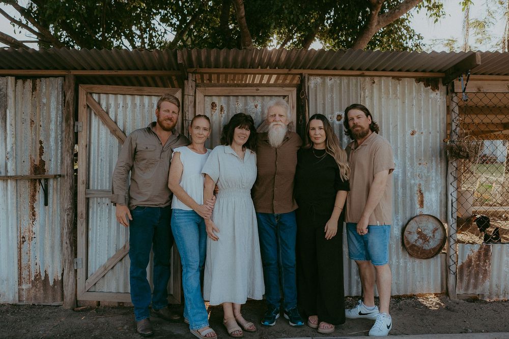 Six adults posing together in front of rustic corrugated iron shed in warm light.