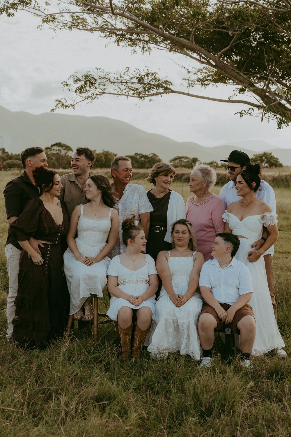 Large extended family group portrait in a grassy paddock with mountains and overcast skies in the background.