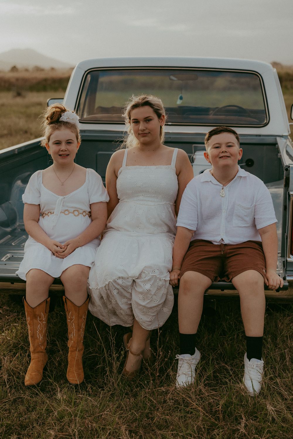 Three siblings sitting together on the back of a vintage ute in a golden grassy field at sunset.