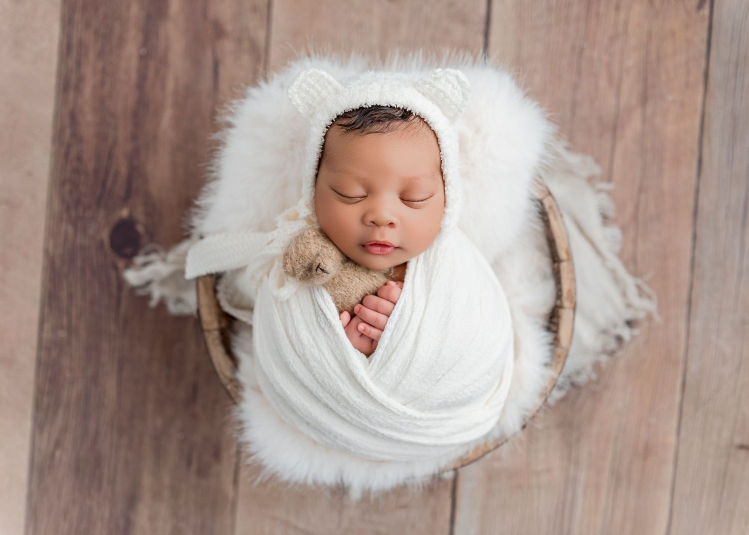 Sleeping newborn wrapped in white fabric posed in rustic wooden bowl.