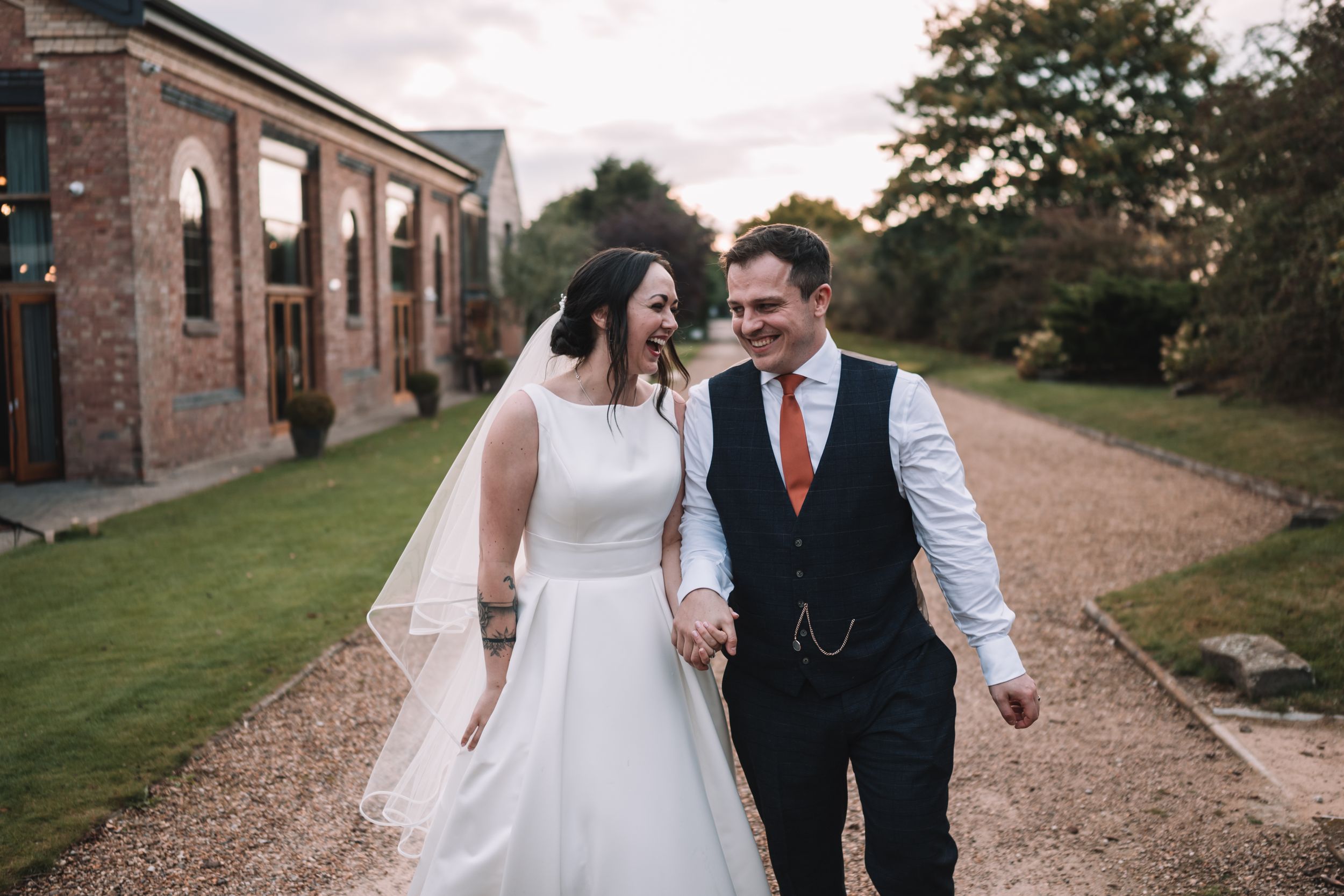A couple in wedding attire walks hand in hand along a gravel path beside the Carriage Hall in Nottingham.