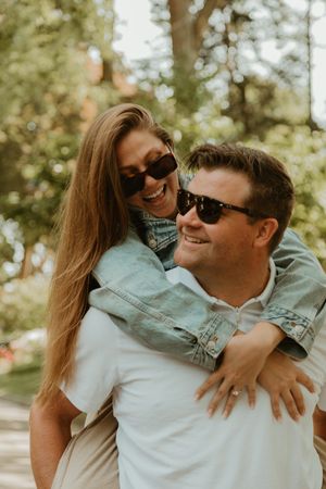 Joyful couple embracing outdoors in sunlit garden while wearing sunglasses and denim jackets.