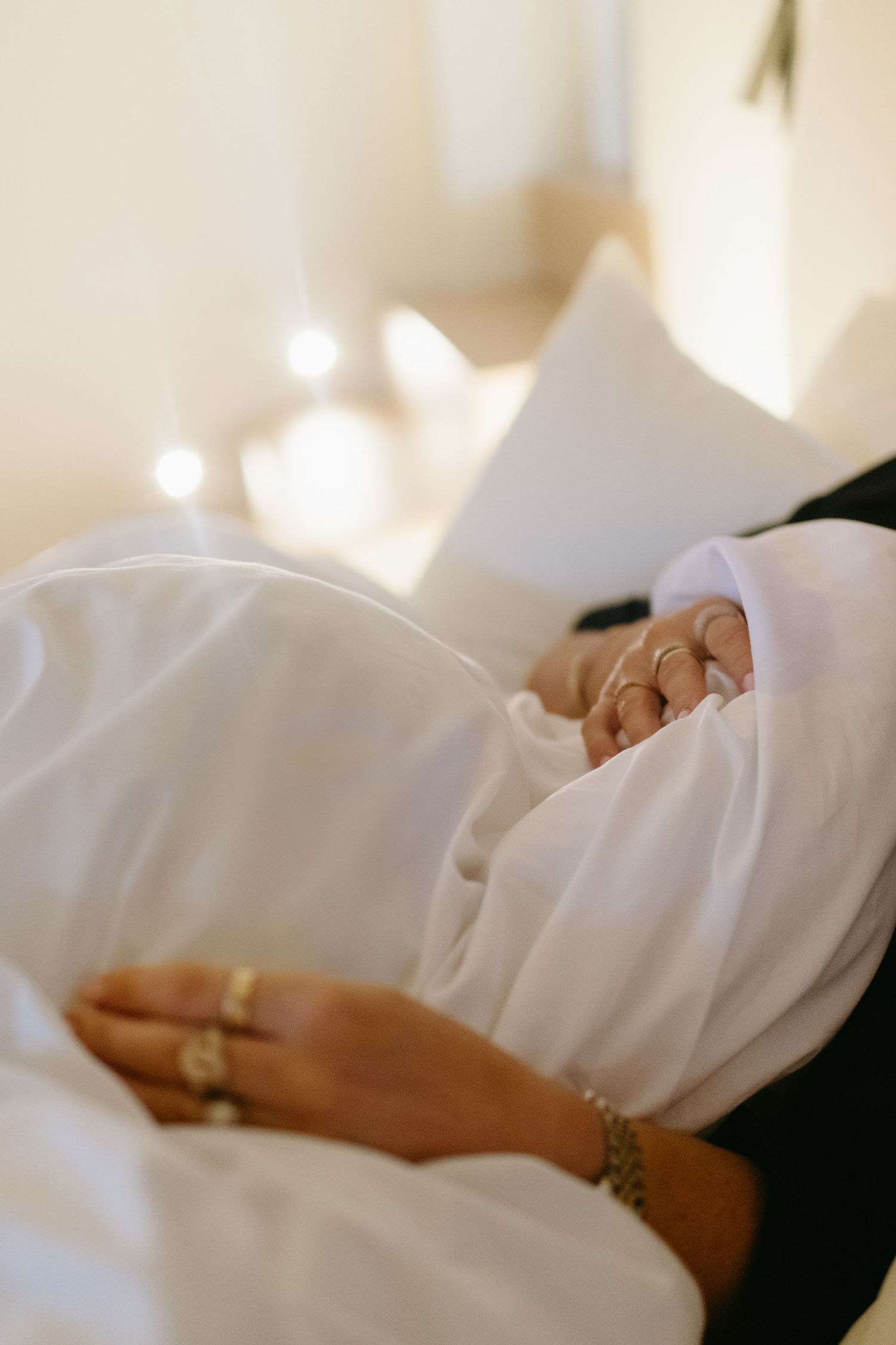detail of woman's hands in bed touching white bed sheets with lights shining in background while being captured by lifestyle photographer