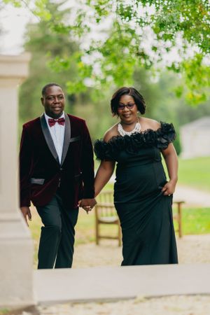A couple walks hand-in-hand in formal evening wear through a garden setting.
