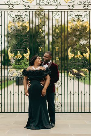 A couple in elegant black formal wear poses in front of ornate gold gates.