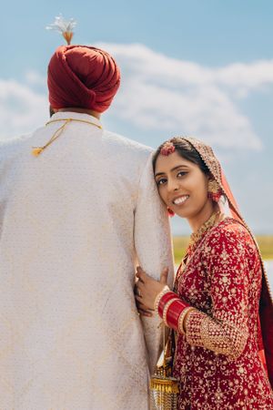 Traditional Sikh bride in red lehenga poses with groom in white sherwani against blue sky background.