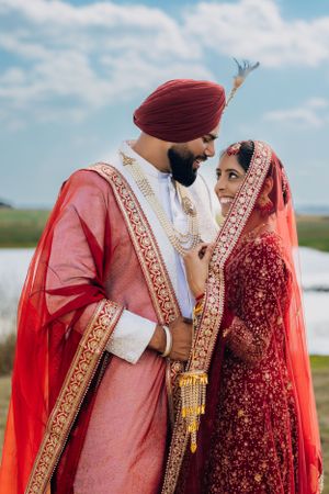 Intimate portrait of couple in ornate red and white traditional wedding outfits against blue sky.