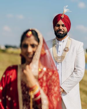 Artistic wedding portrait showing groom in white sherwani with red turban and bride in traditional red dress.